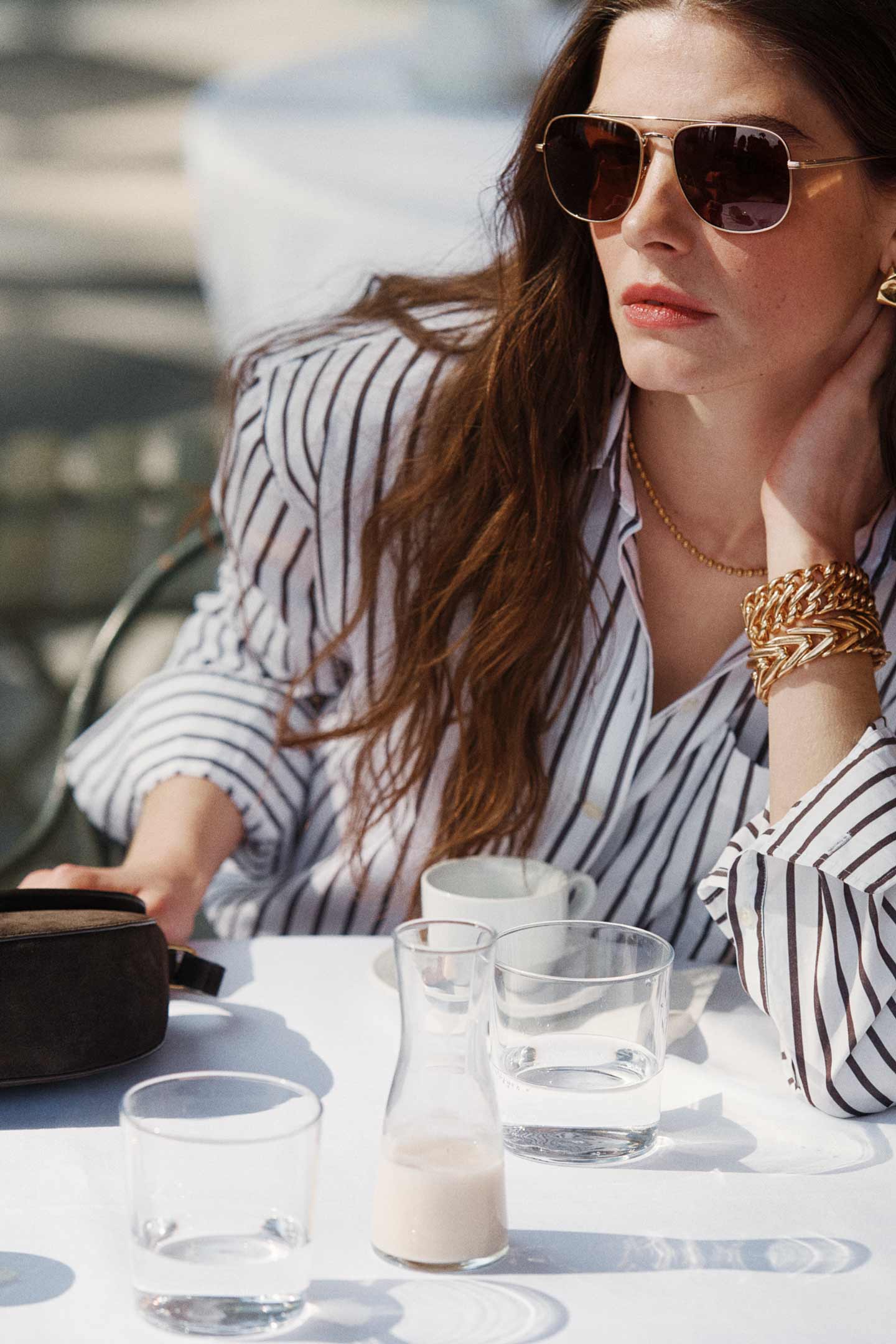 Model styled in a GANT striped shirt with accessories seated at an outdoor table.