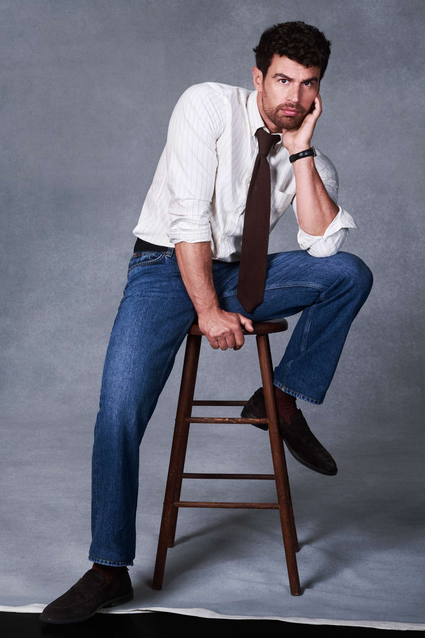 Actor Theo James styled in a Gant button-down shirt, tie and jeans while seated on a stool in a studio setting.