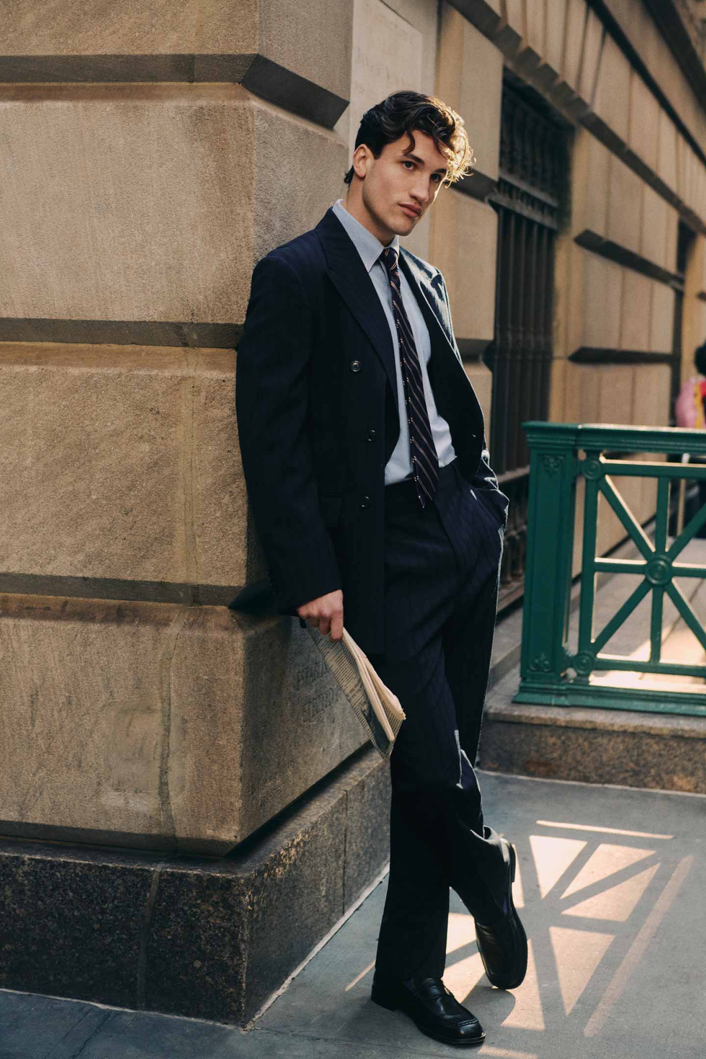 Person walking in a dark blue Gant blazer and pants with a striped tie and dress shirt in an urban street setting.