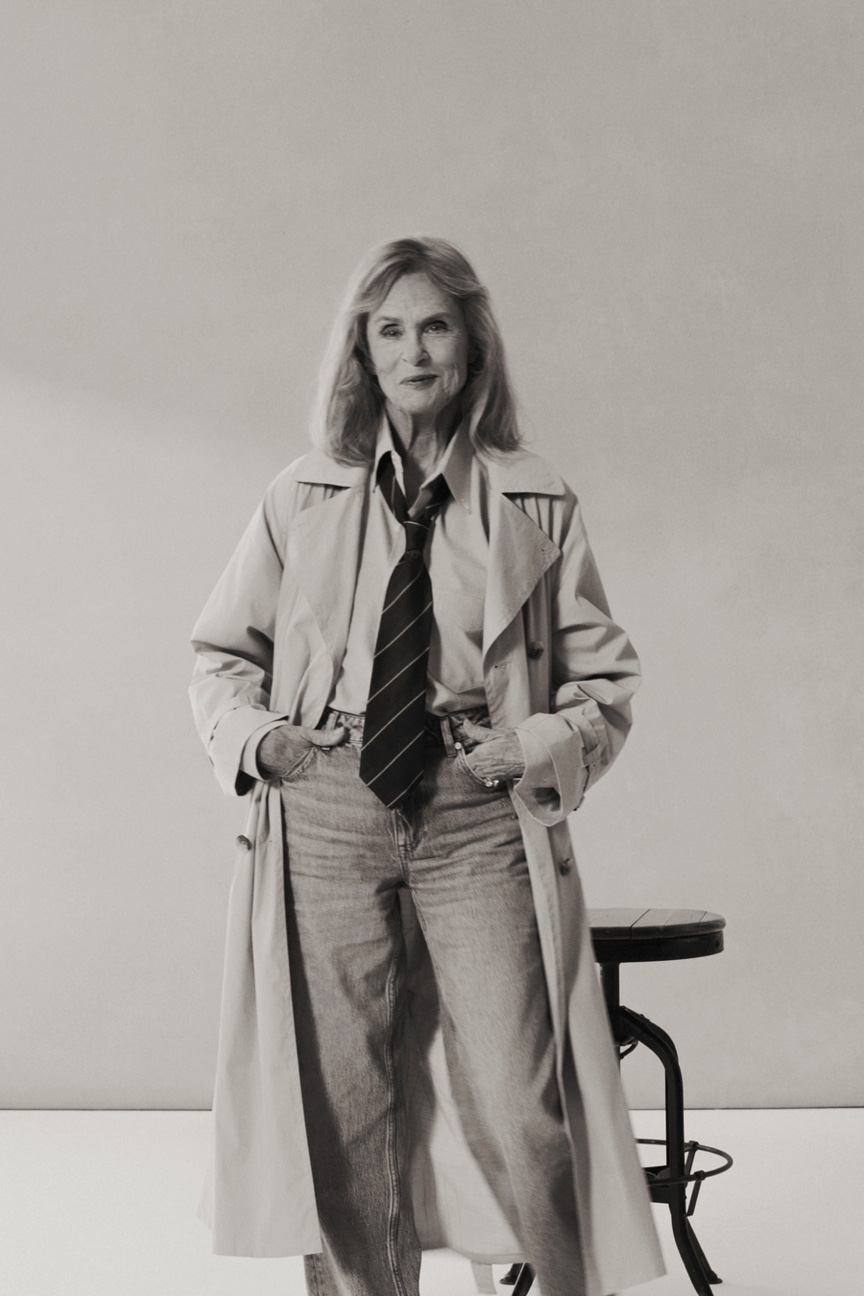 Lauren Hutton wearing a Gant trench coat, shirt, tie and jeans standing in a studio setting