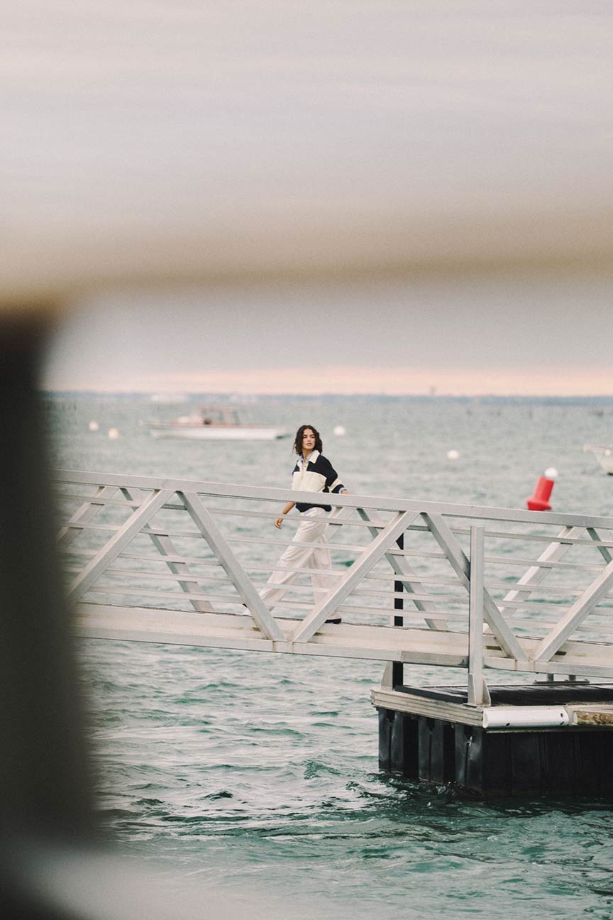 Donna che cammina su un molo indossando una maglia da rugby Gant a blocchi di colore e pantaloni bianchi.