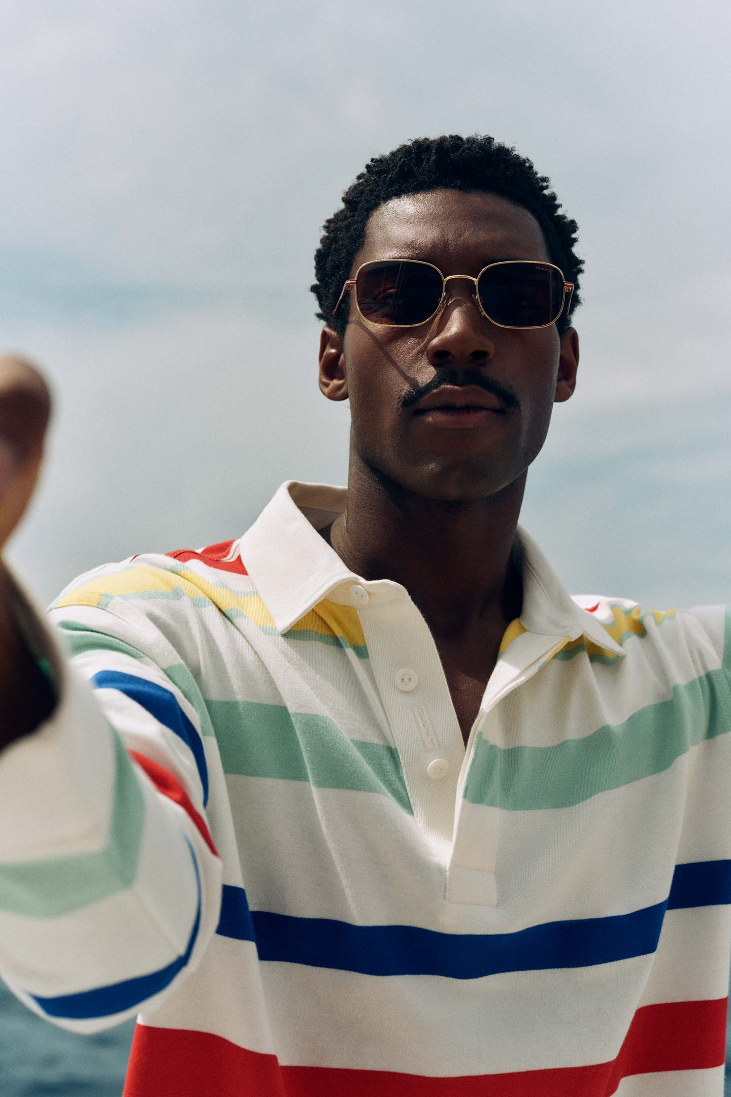 Man wearing a Gant striped rugby shirt standing near the sea with arms extended in an outdoor setting