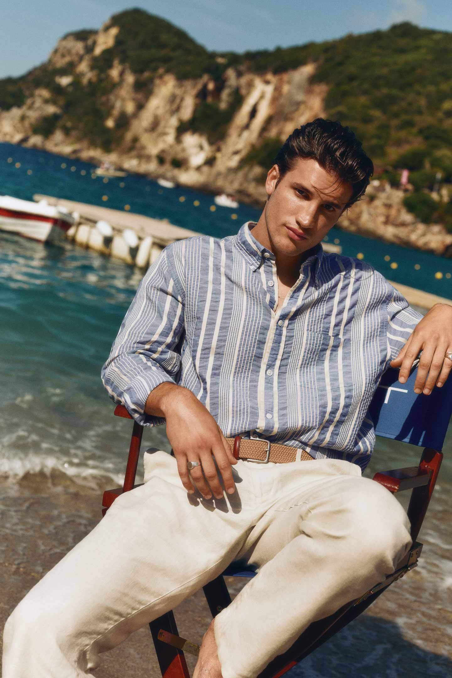 Man wearing a Gant blue‑and‑white striped linen shirt with beige trousers sitting on a chair by the water.