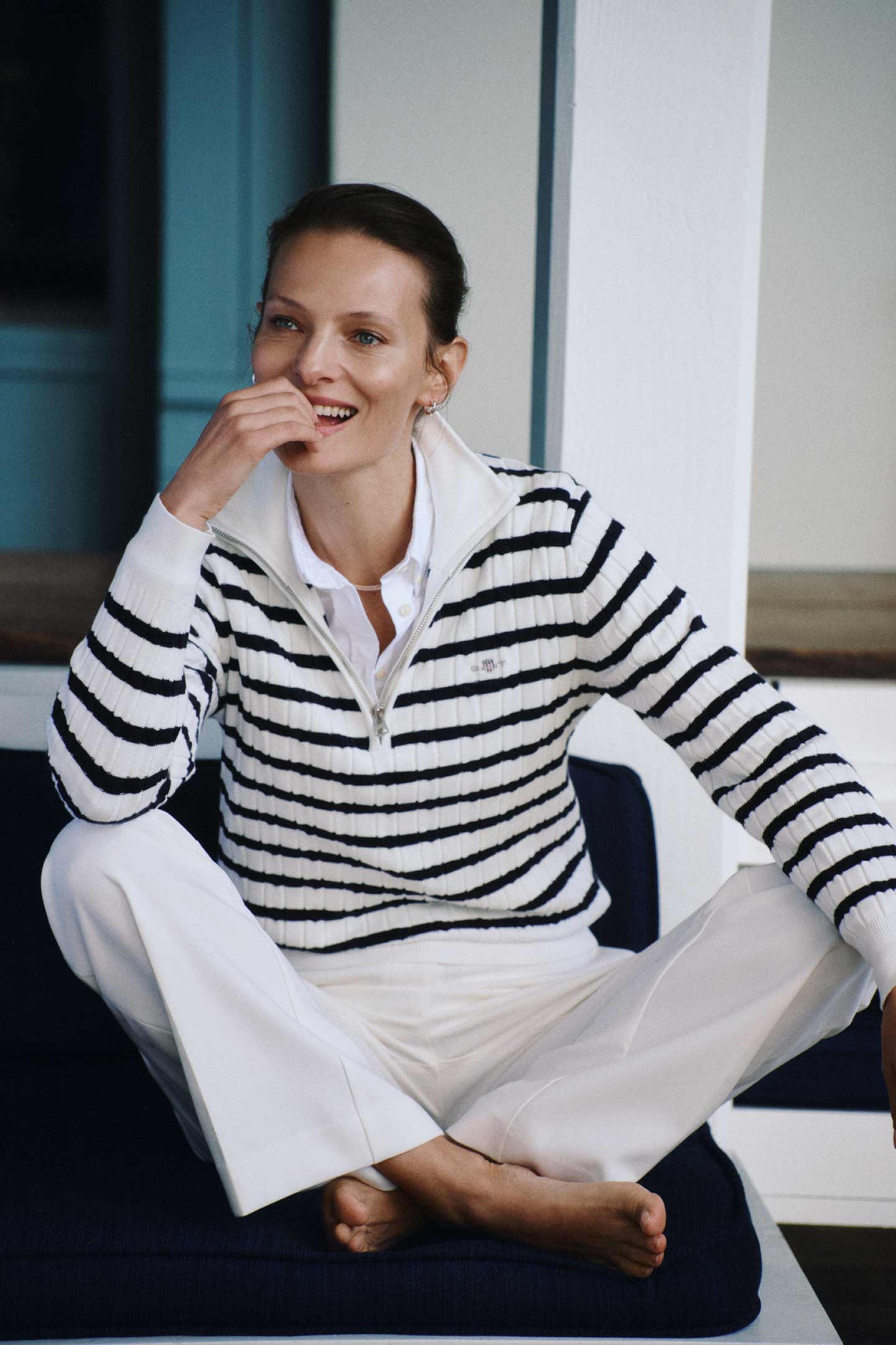 Woman wearing a Gant striped half‑zip sweater over a white shirt with white trousers while seated indoors
