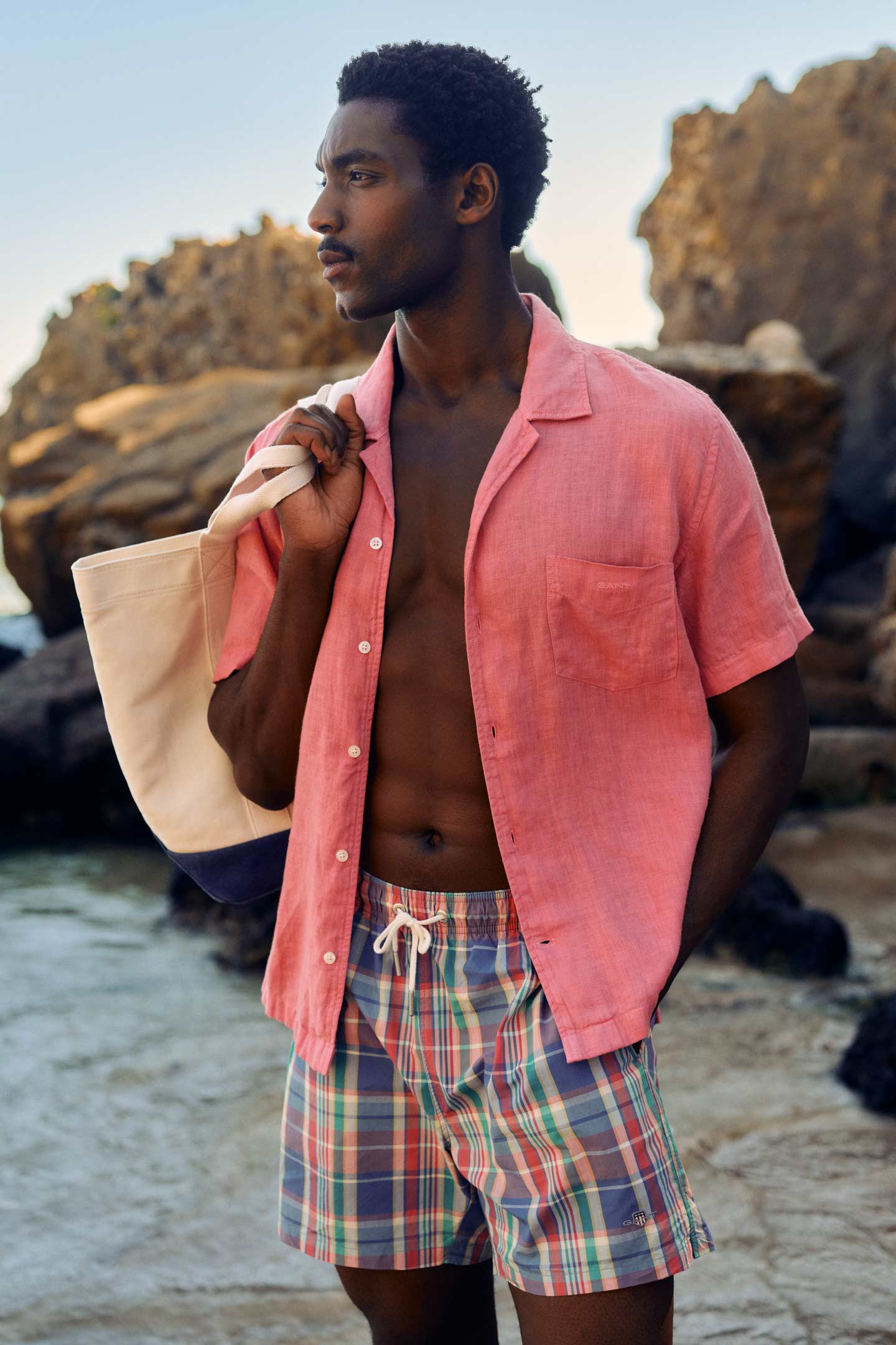 Man standing by rocky shoreline wearing a pink short‑sleeve linen shirt over plaid swim shorts, holding a two‑tone canvas tote bag.