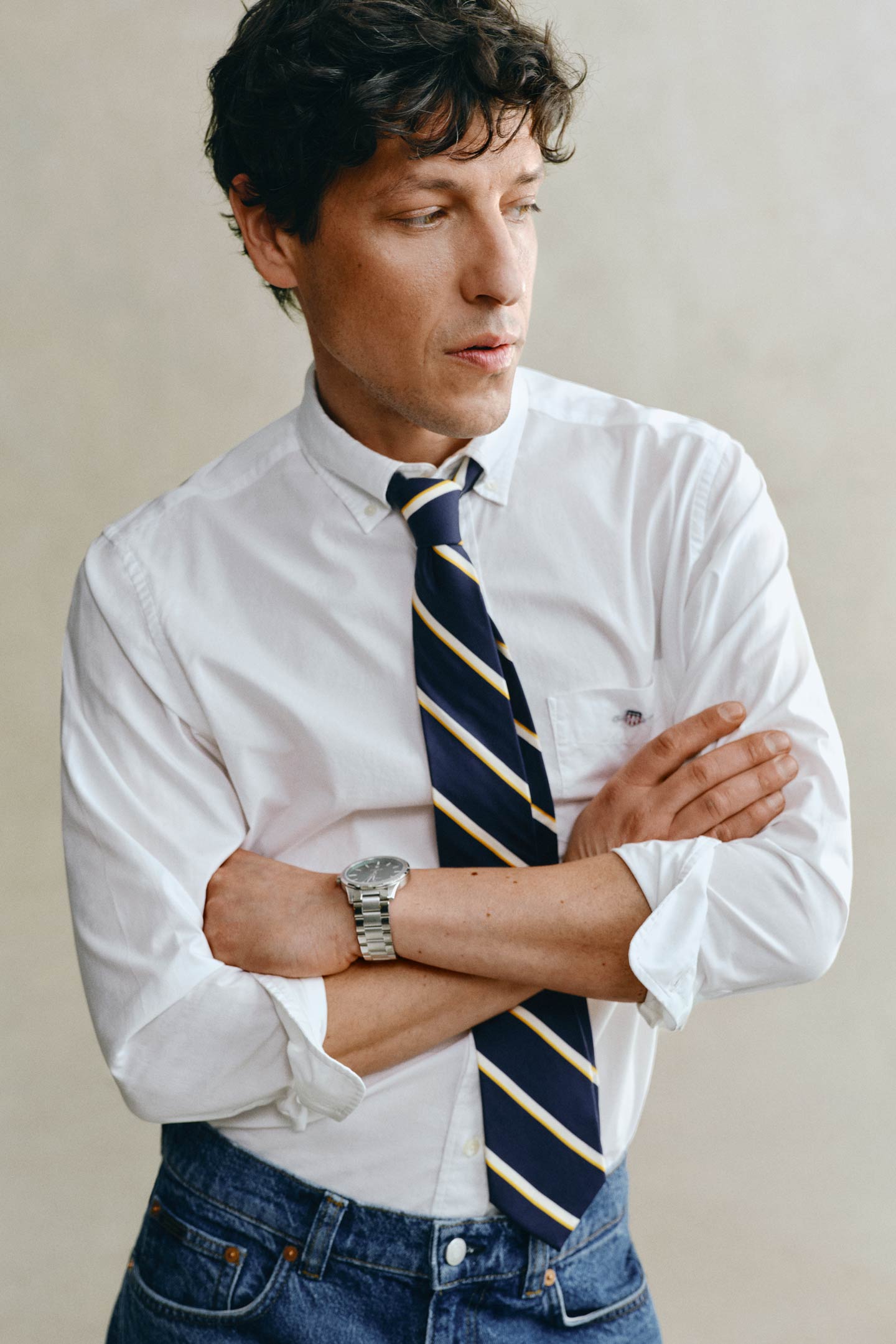 Model styled in a GANT white shirt, tie and blue jeans with arms crossed in front of a grey backdrop.