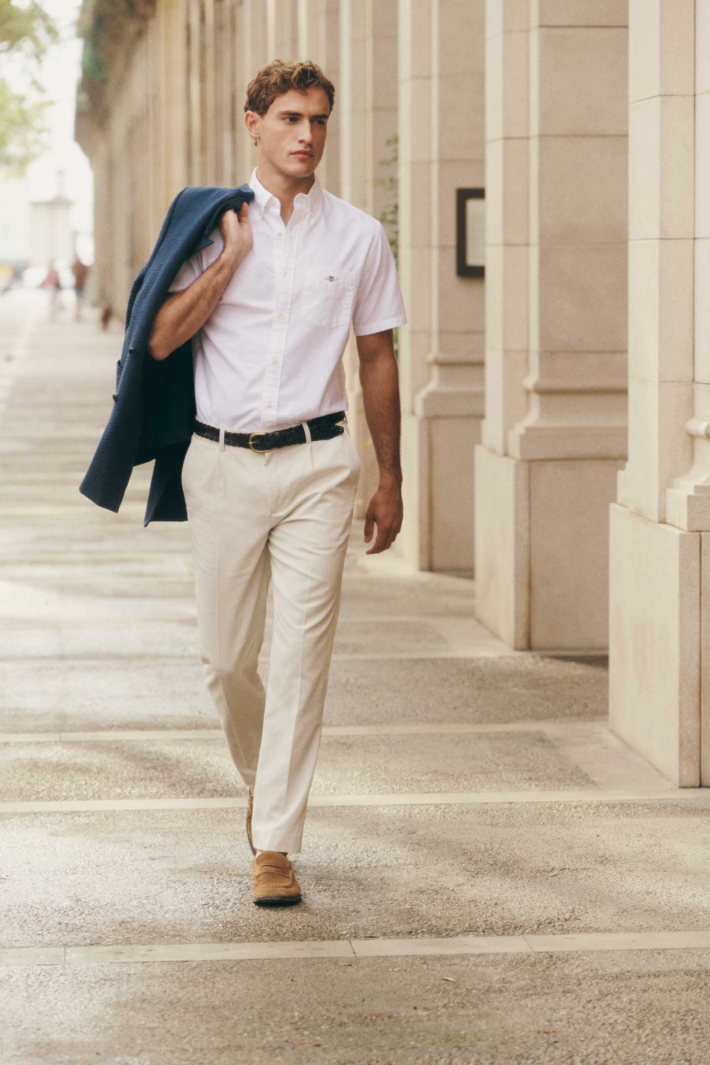 Man wearing a Gant white short-sleeve shirt and light trousers while walking with a blazer.