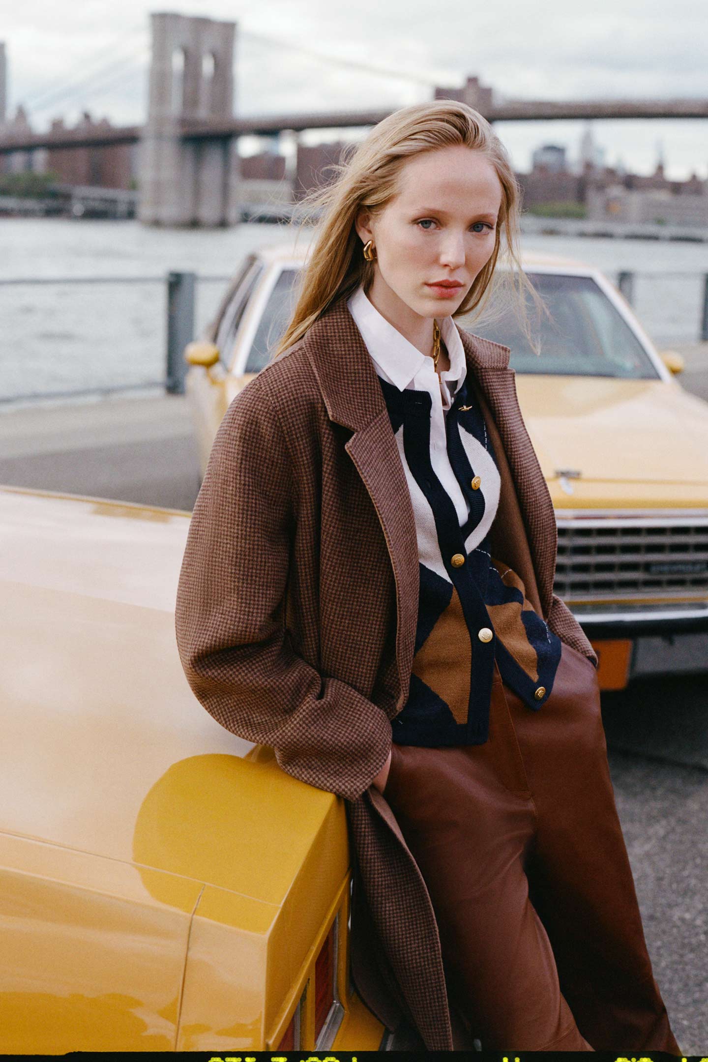 Model styled in a Gant checked overcoat worn over an argyle knit cardigan, shirt and leather pants, leaning against a yellow taxi.