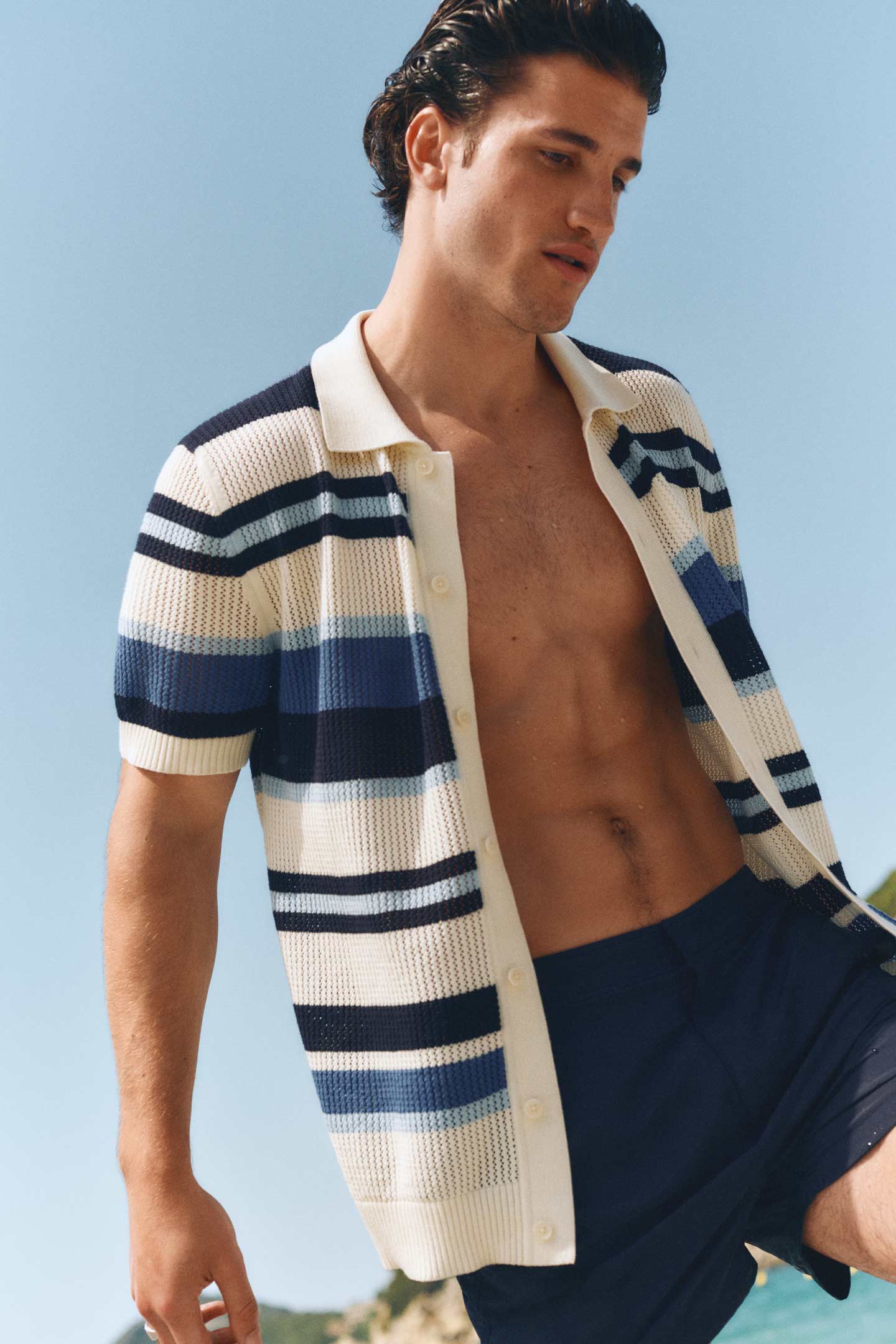 Man wearing a Gant striped short‑sleeve knit shirt and navy swim shorts while standing outdoors near the shoreline