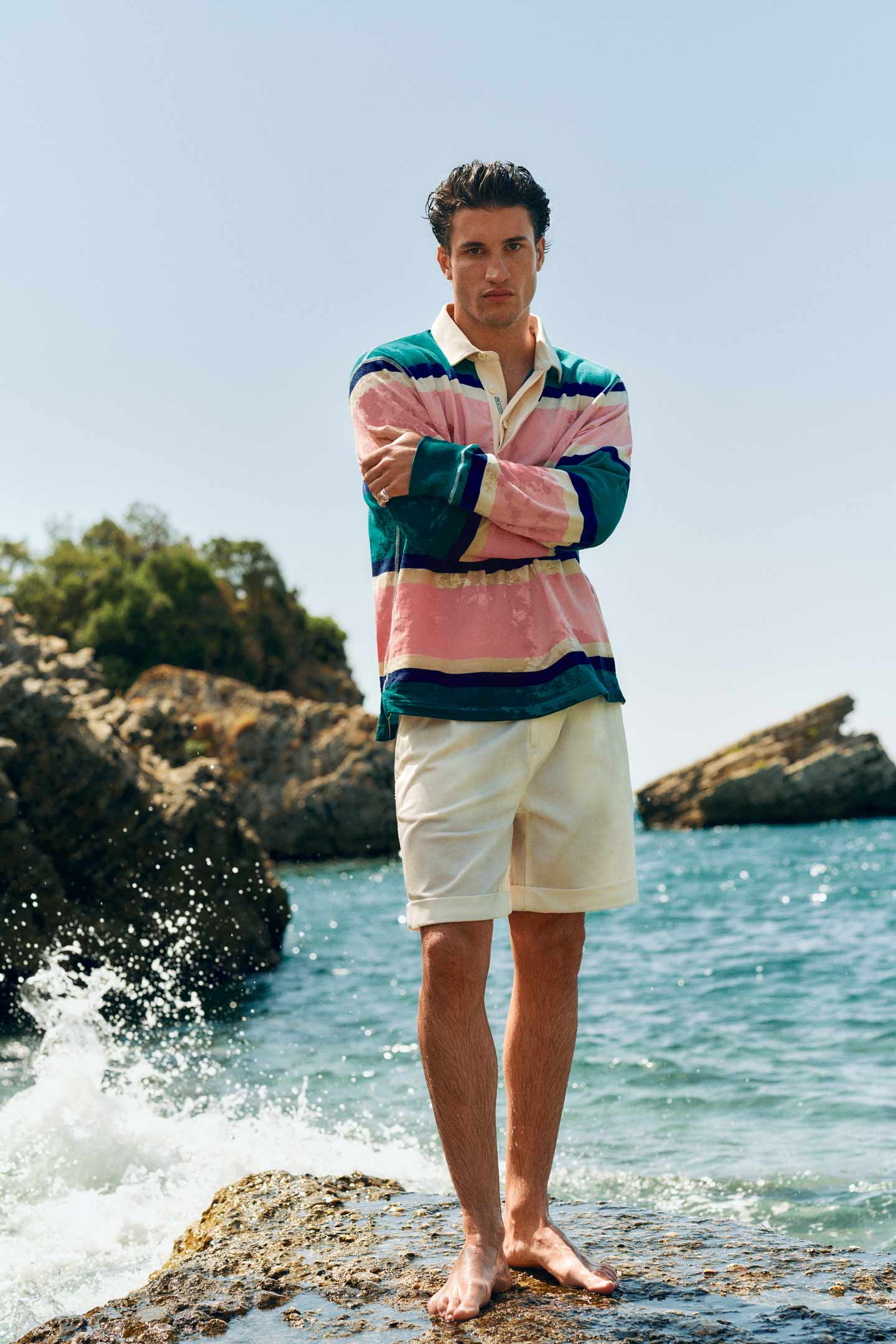 Man wearing a Gant multicolour striped rugby shirt with beige shorts by the shoreline.