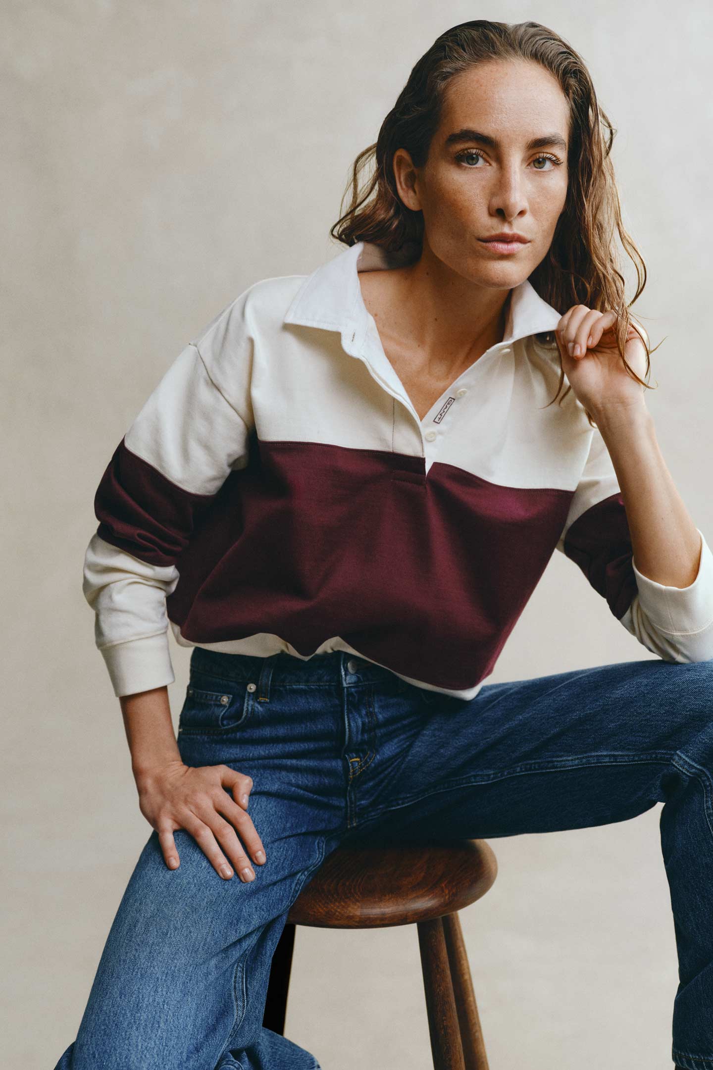 Model styled in a GANT color-blocked rugger layered over a shirt with jeans sitting in front of a grey backdrop.
