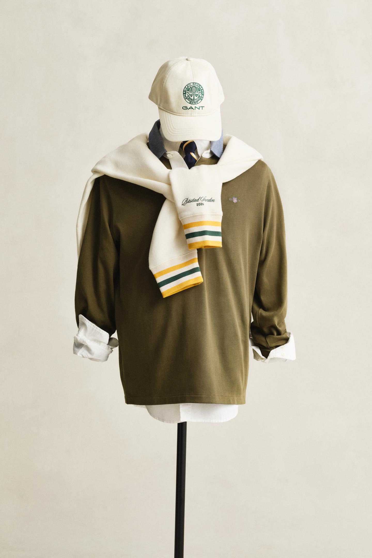Gant rugby shirt layered over a collared shirt with a sweater tied at the shoulders and a cap displayed on a mannequin.