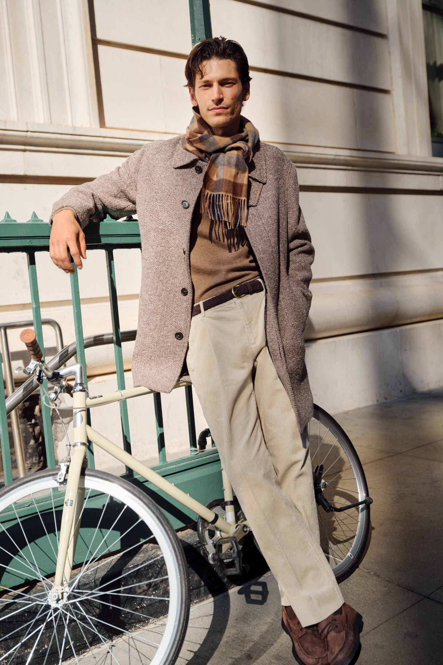 Model styled in a Gant herringbone coat over a knit sweater, checked scarf and corduroy pants beside a bicycle.