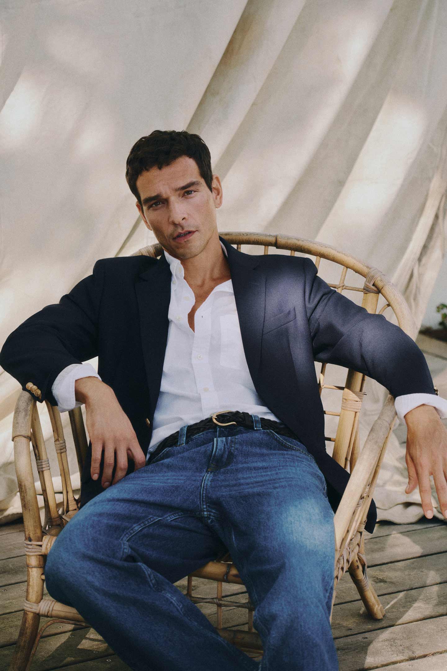 A man wearing a Gant white button‑down shirt and a navy blazer paired with blue jeans, seated in a woven chair.