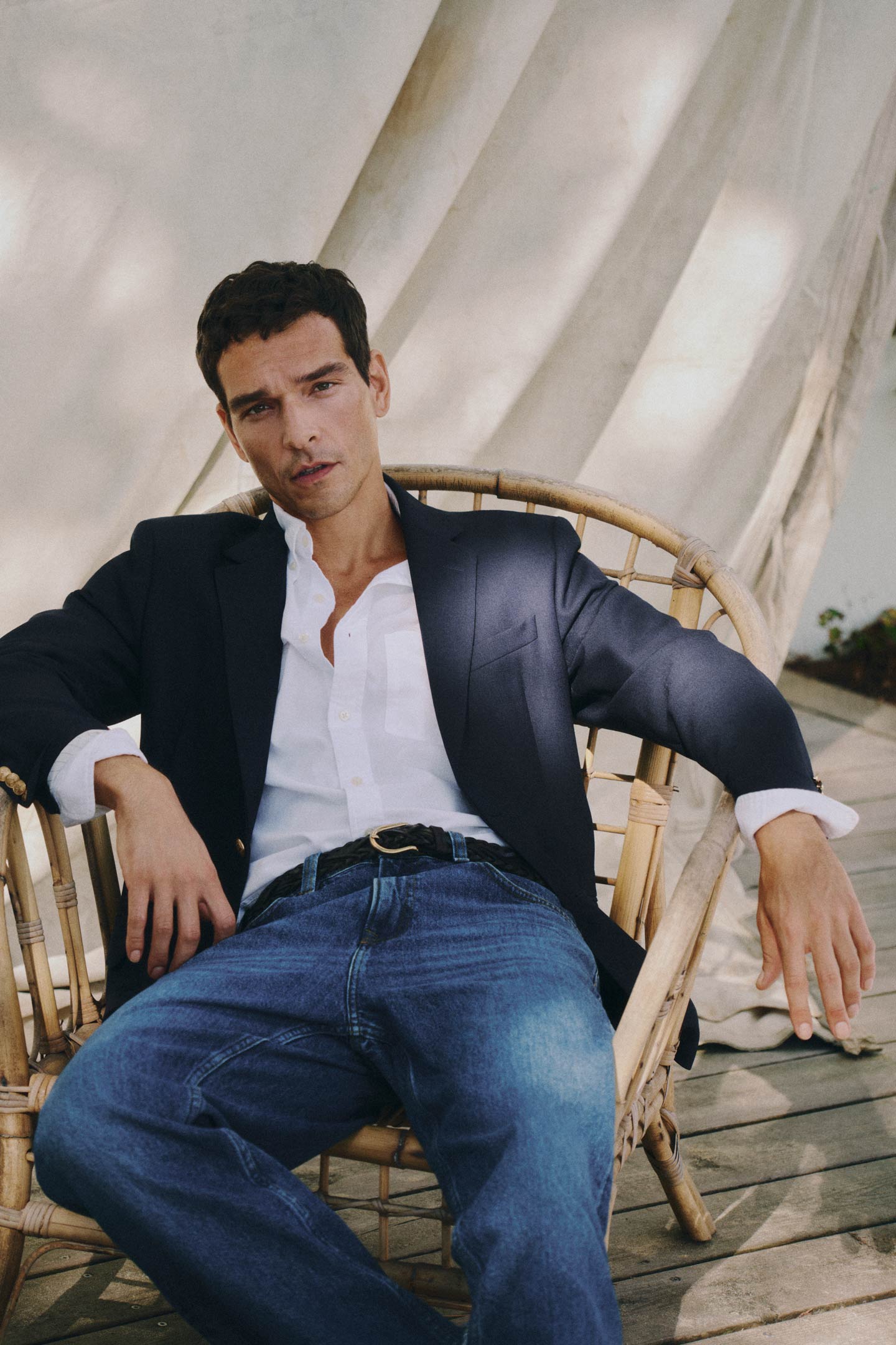 A man wearing a Gant white button‑down shirt and a navy blazer paired with blue jeans, seated in a woven chair.