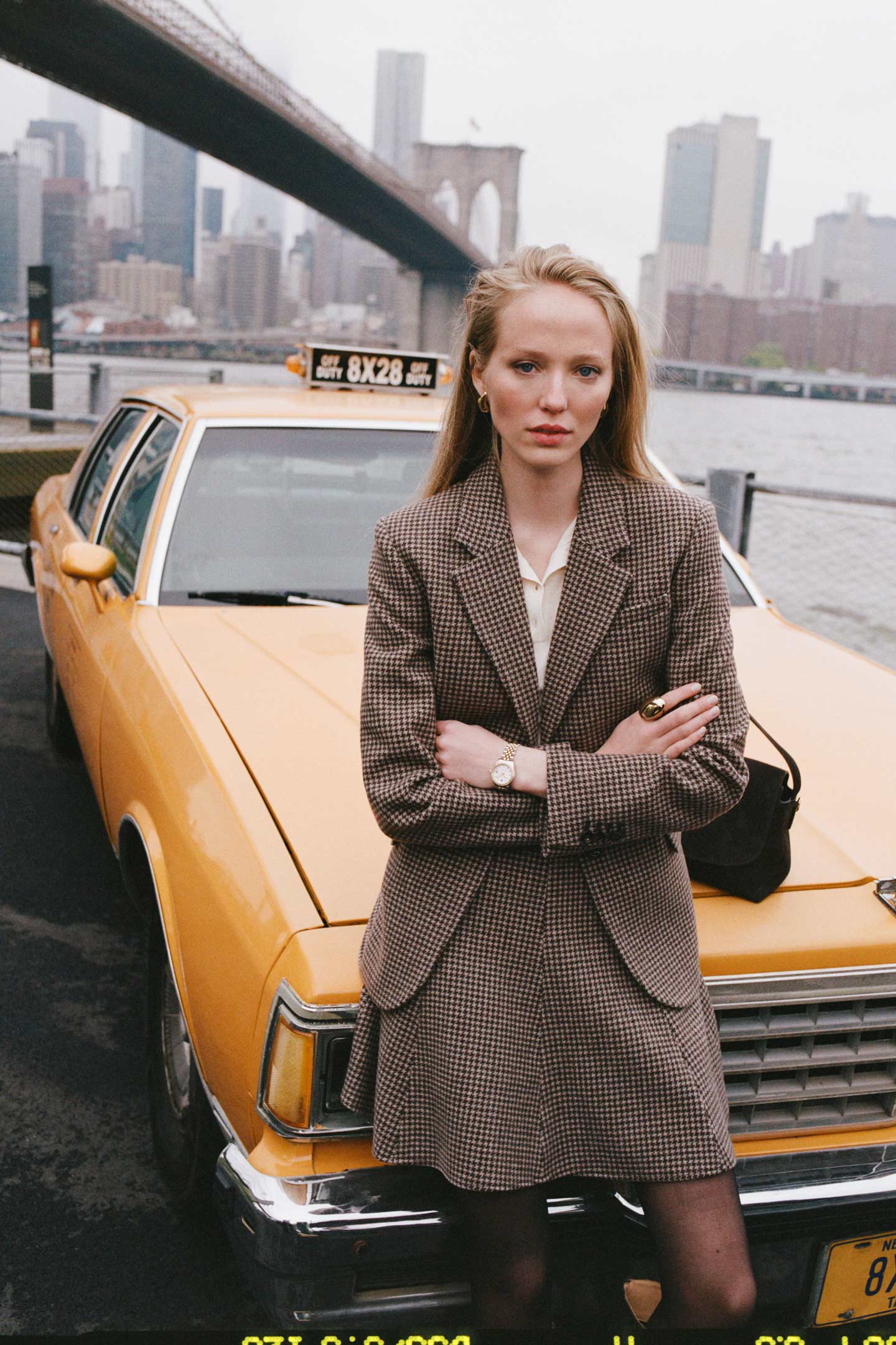 Model styled in a Gant checked blazer and matching skirt styled with a blouse and shoulder bag posing seated on the bonet of a yellow taxi.