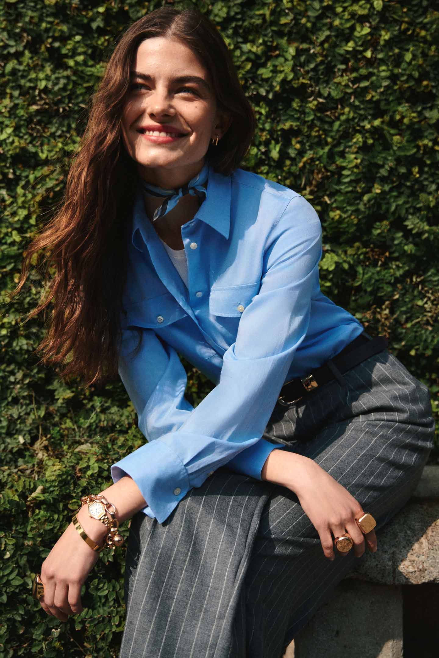 Model styled in a GANT blue shirt paired with striped tailored pants sitting in front of greenery.