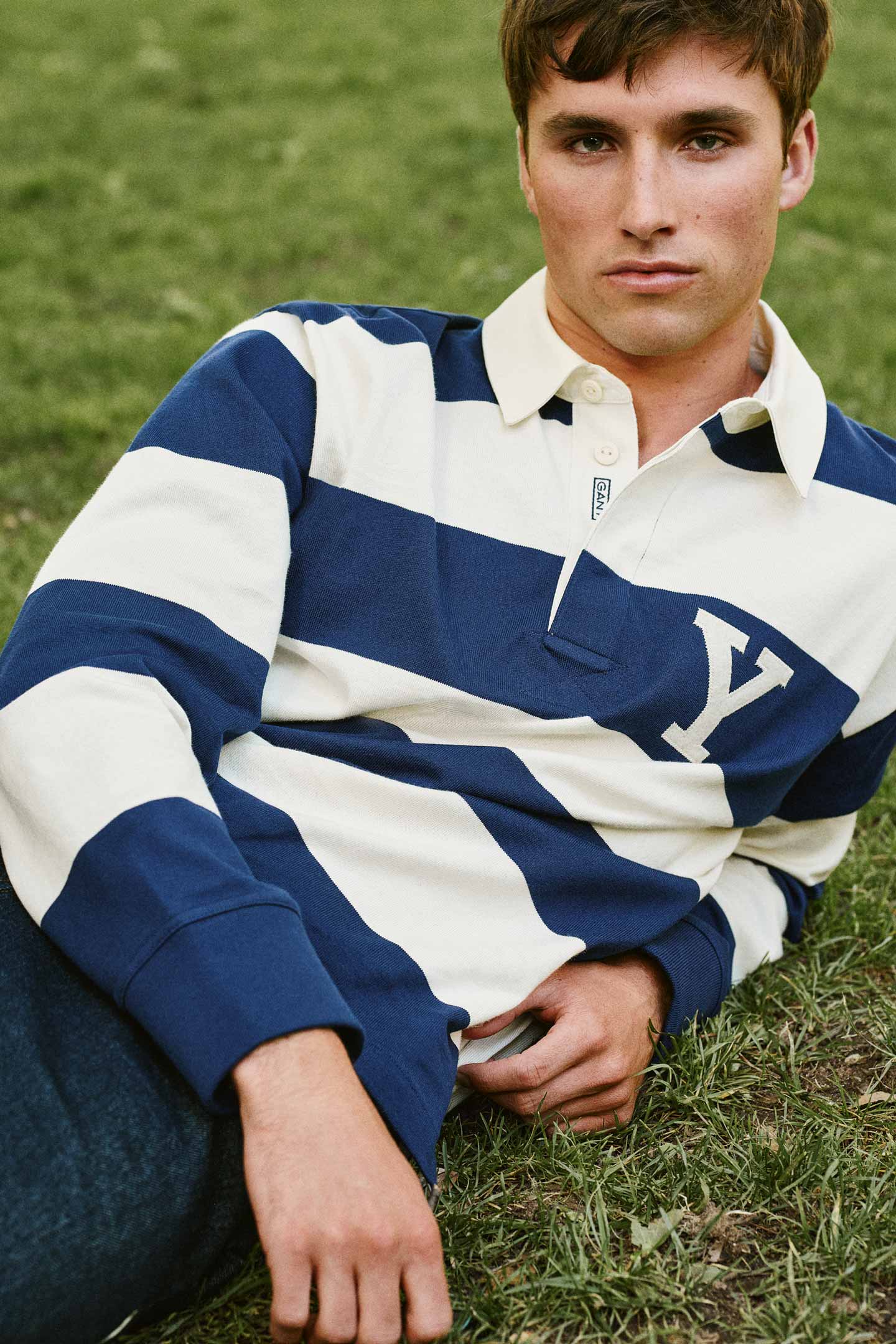 Model styled in a GANT striped rugger and pants resting on grass outdoors.