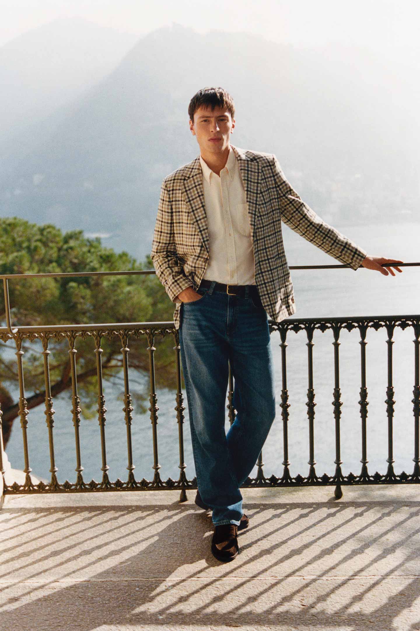 Model styled in a GANT checked blazer, shirt and jeans standing on a terrace overlooking a lake.