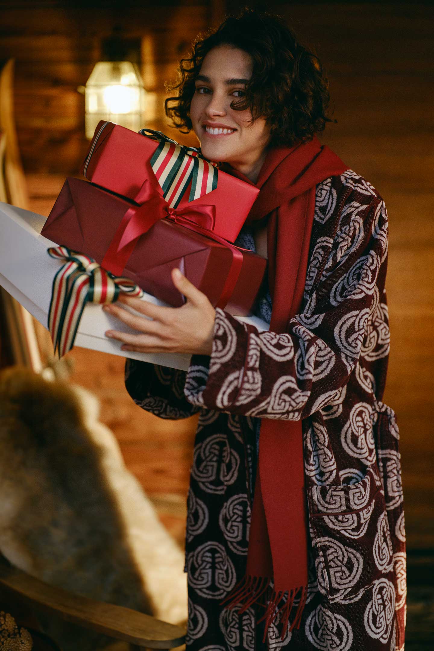 Model styled in a Gant patterned robe layered with a scarf, holding wrapped gifts in a lodge interior.