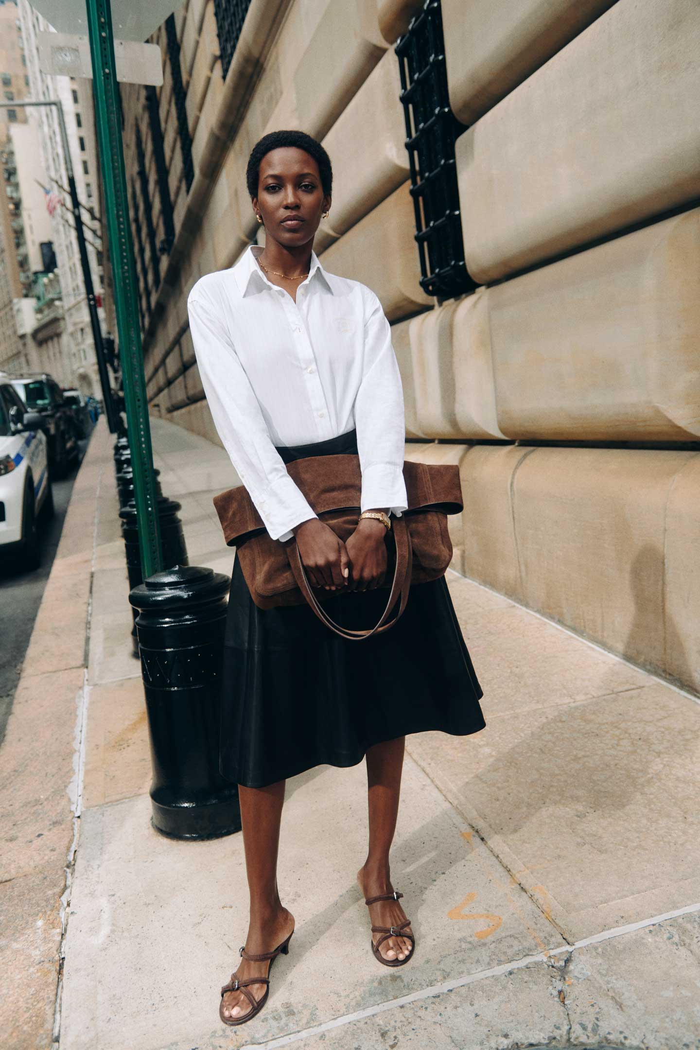 Person wearing a Gant white shirt with a black skirt and carrying a brown leather bag on a city sidewalk.
