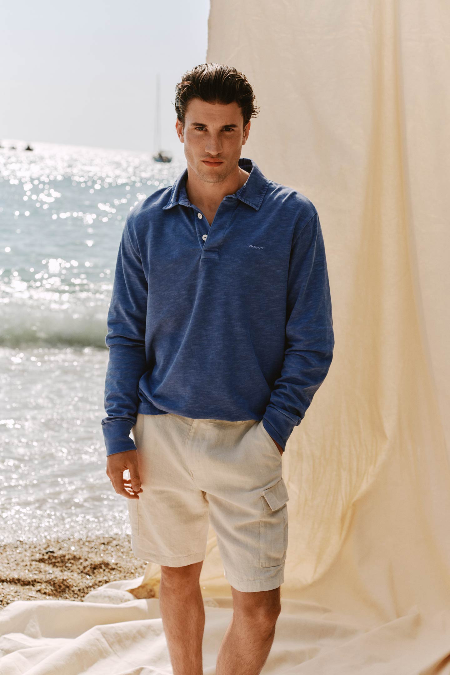 Man standing by the shore wearing a Gant blue long-sleeve polo and beige shorts.