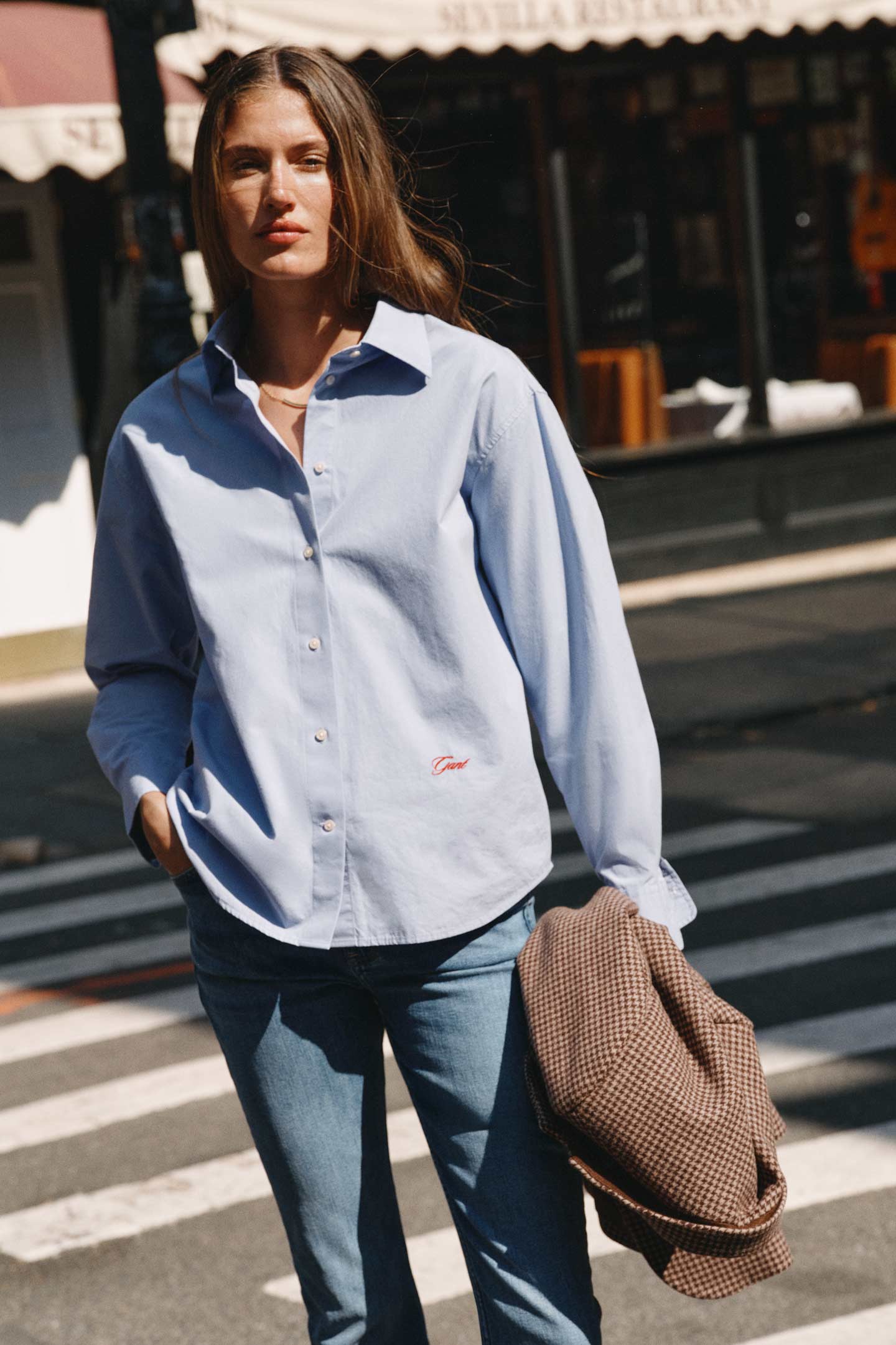 Model styled in a light blue Gant cotton shirt paired with jeans, carrying a checked blazer while crossing a city street.