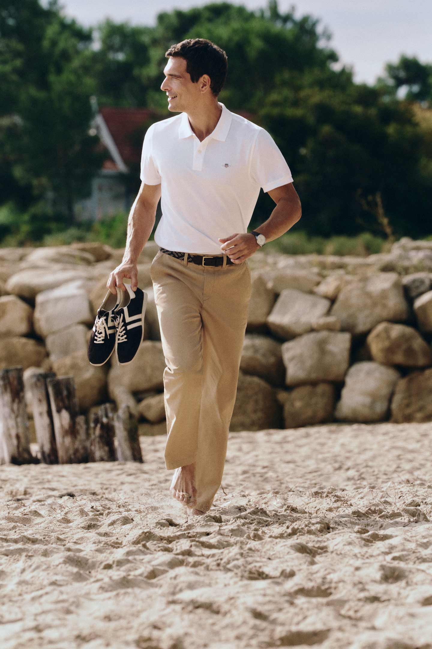 Person wearing a white short‑sleeve polo shirt paired with beige trousers and a dark belt, walking barefoot on a sandy beach while holding a pair of black sneakers.