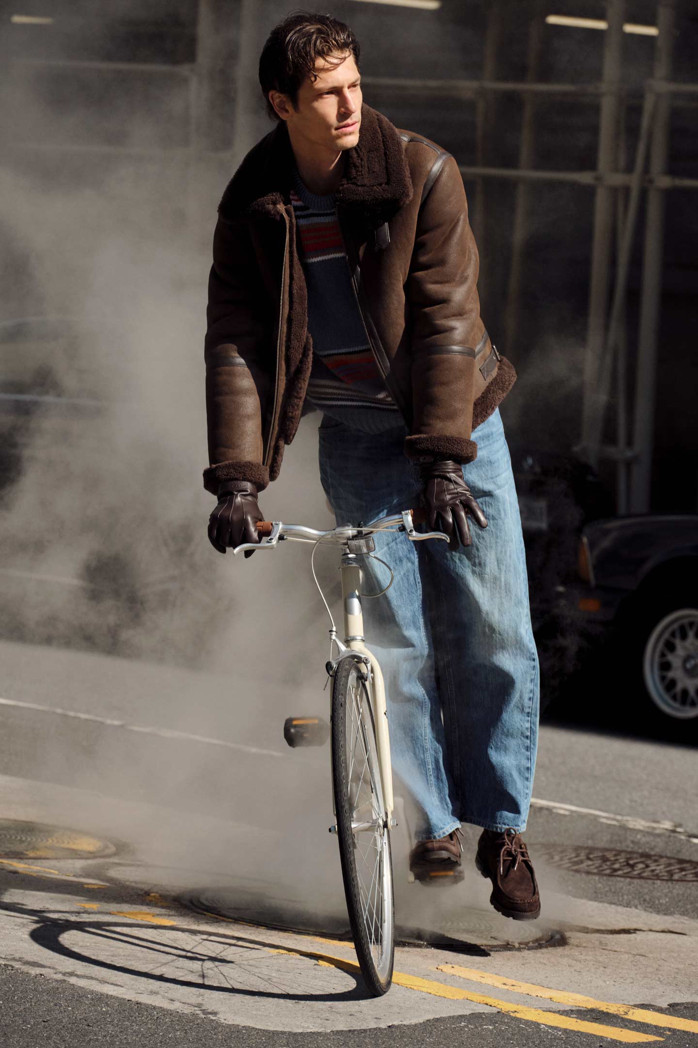 Model styled in a Gant shearling jacket worn while riding a bicycle on a city street, paired with jeans and gloves.