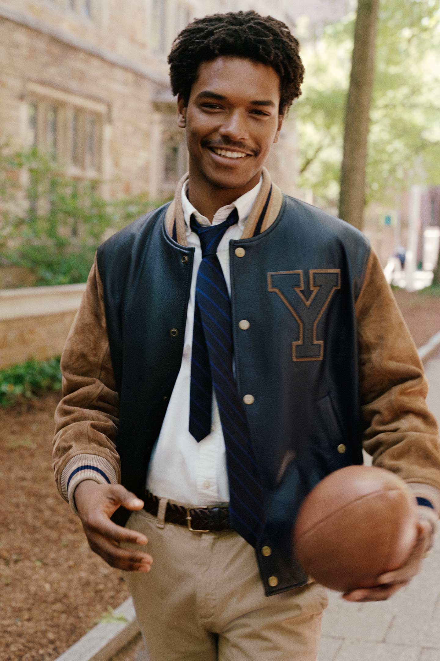 Model styled in a GANT varsity jacket with shirt and tie holding a basketball outdoors.