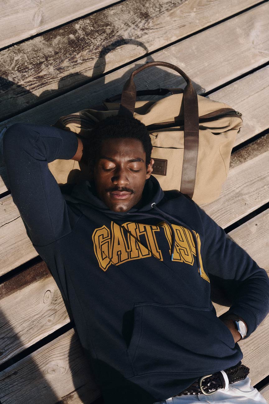Uomo sdraiato su un ponte di legno che indossa una felpa con cappuccio con logo Gant accanto a una borsa da weekend in tela.