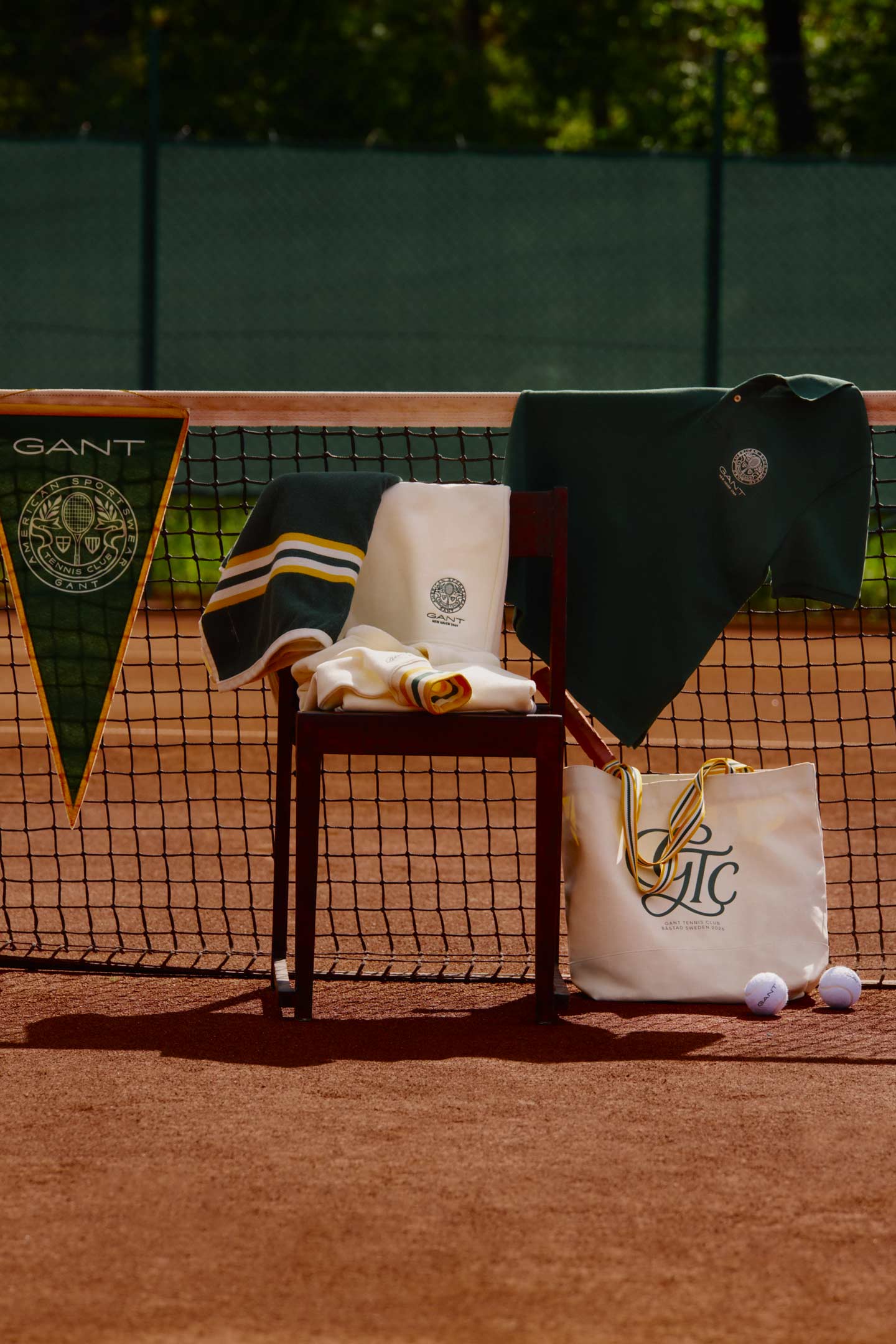 Gant tennis clothing and knitwear displayed on a chair with a tote bag and tennis balls beside a court net outdoors.