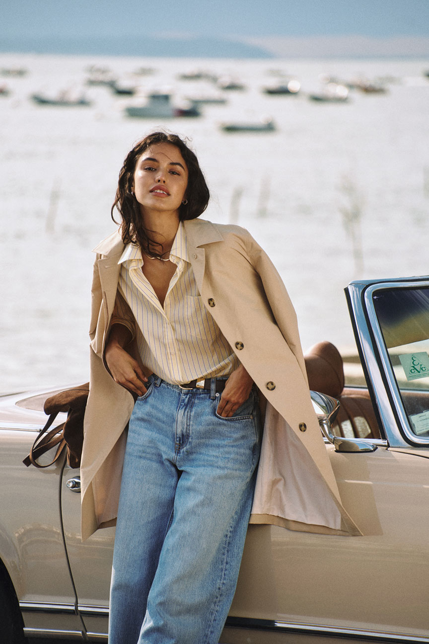 Donna appoggiata a un'auto che indossa un trench Gant, una camicia a righe e jeans a gamba larga vicino all'acqua.