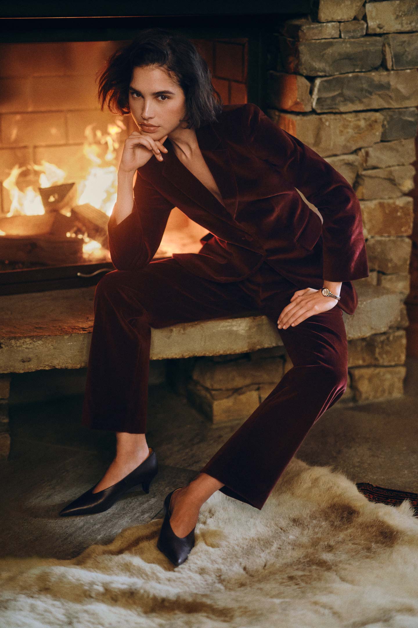 Model styled in a Gant velvet trouser suit paired with heeled shoes, seated beside a lit stone fireplace.