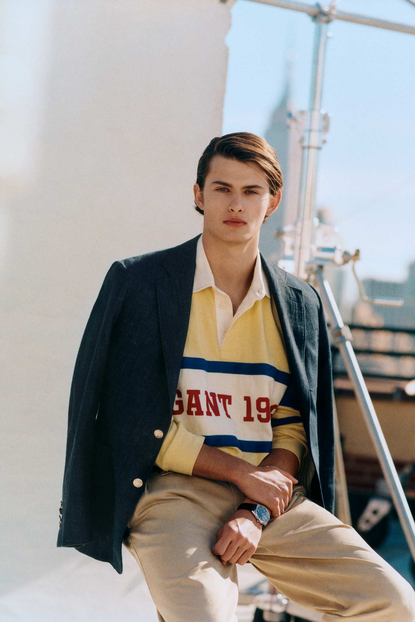 Man wearing a Gant yellow rugby shirt with logo, navy blazer, and beige trousers outdoors.