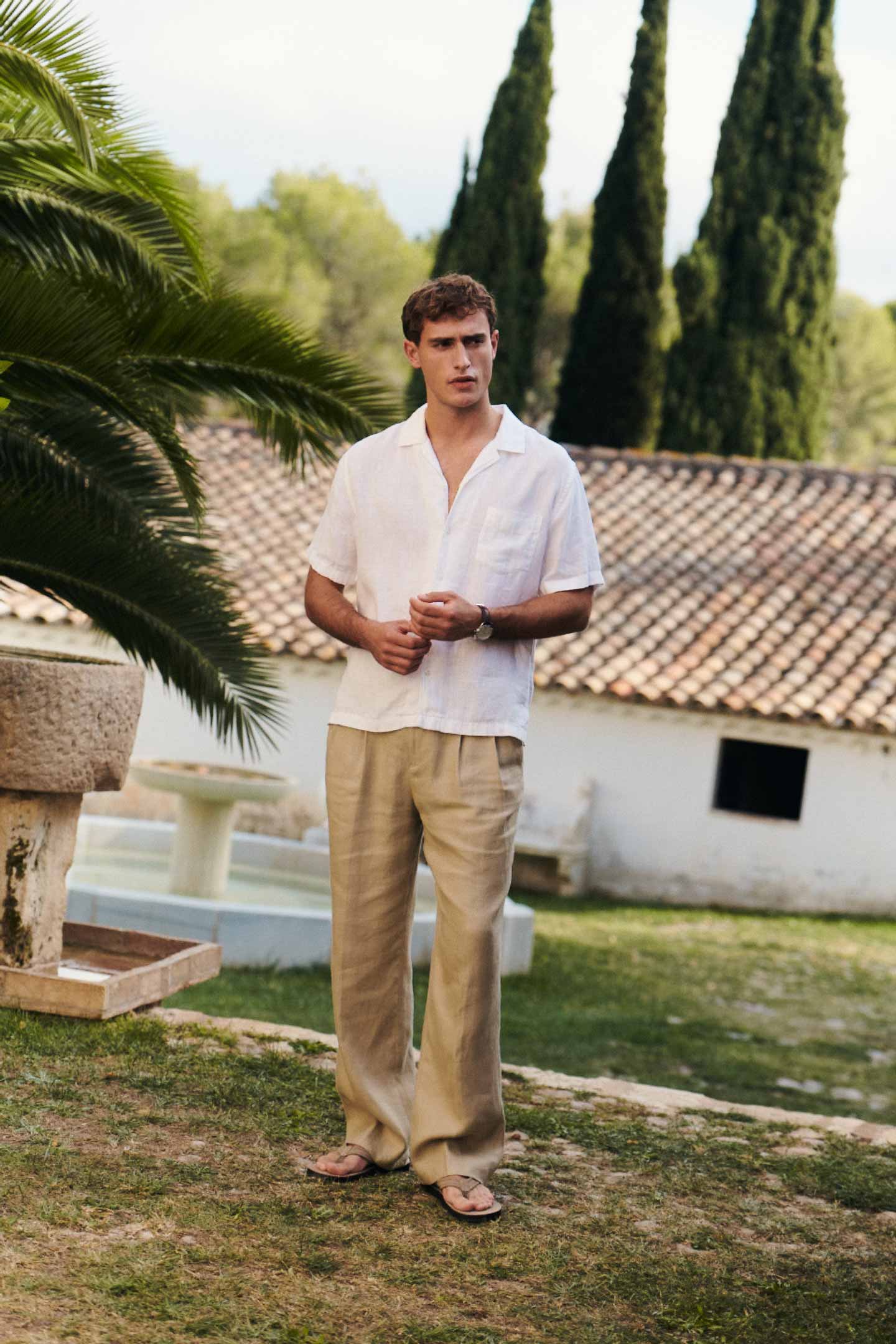 Man wearing a Gant white linen shirt and beige linen trousers outdoors near a tiled roof building.