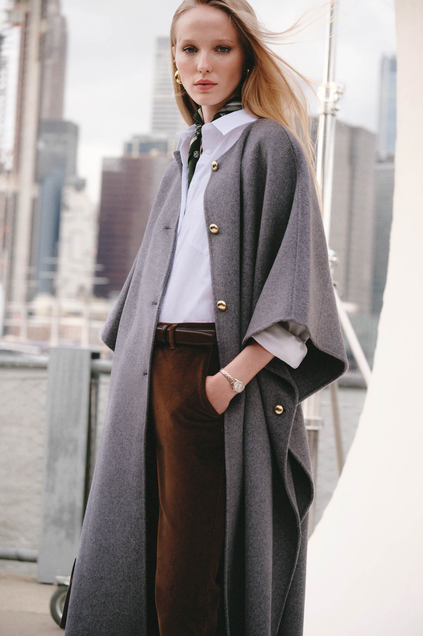 Model styled in a Gant cape-style coat layered over a shirt and pants, finished with a neck scarf against a city backdrop.