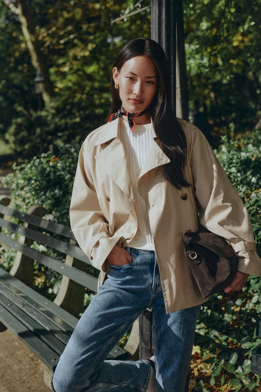  A person wearing classic blue GANT jeans paired with a lightweight beige trench jacket and white top, standing by a park bench in a sunlit garden setting.