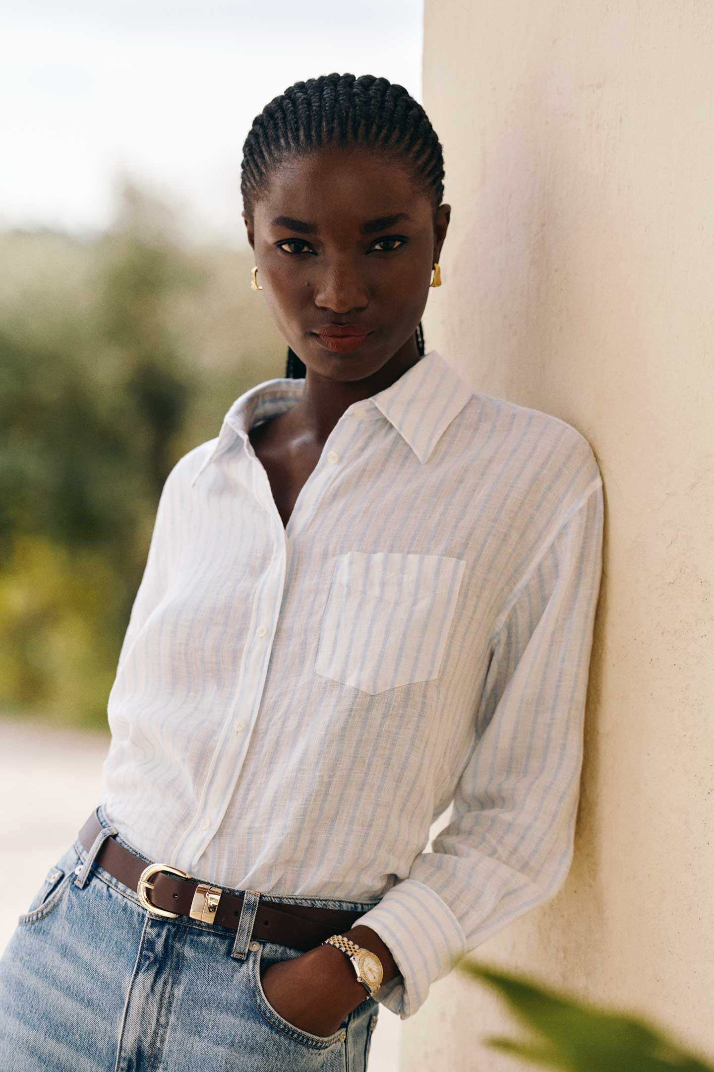 Woman wearing a Gant light striped button‑down shirt with blue jeans and a brown belt while leaning against a wall outdoors