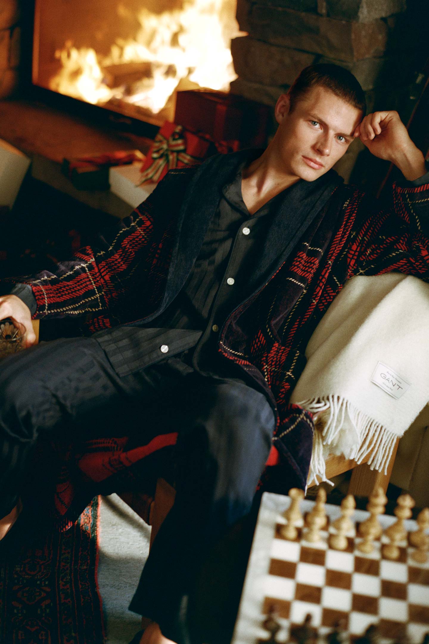 Model styled in a Gant checked robe layered over satin pyjamas, seated beside a fireplace.