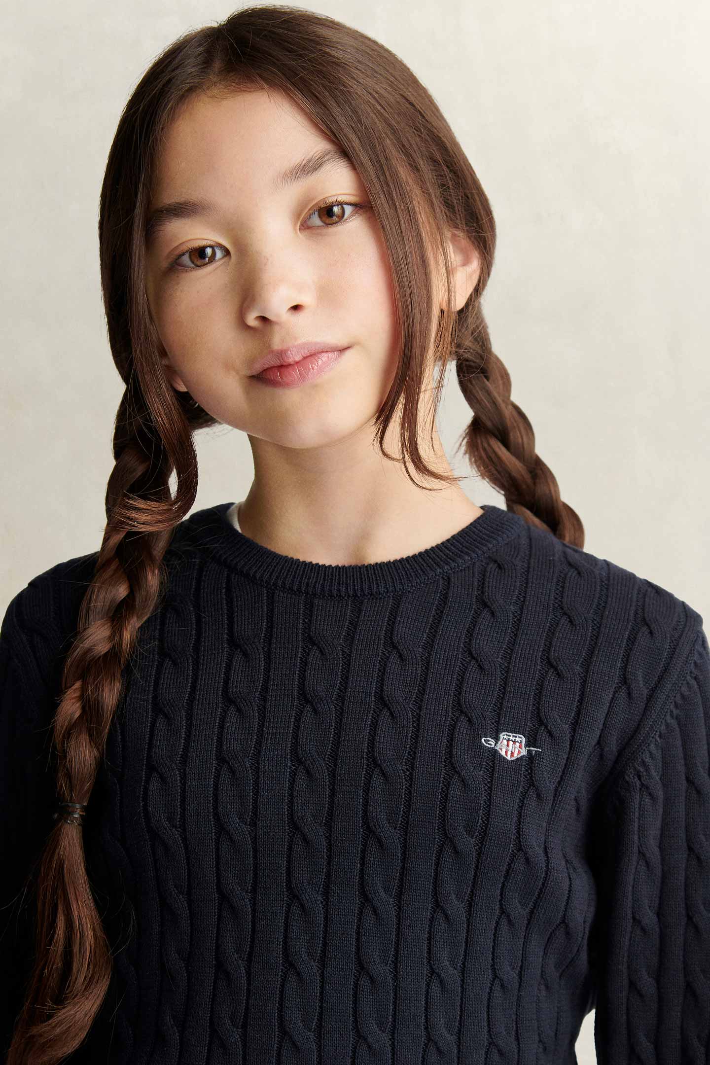 Child wearing a Gant cable-knit sweater in navy blue with embroidered shield logo.