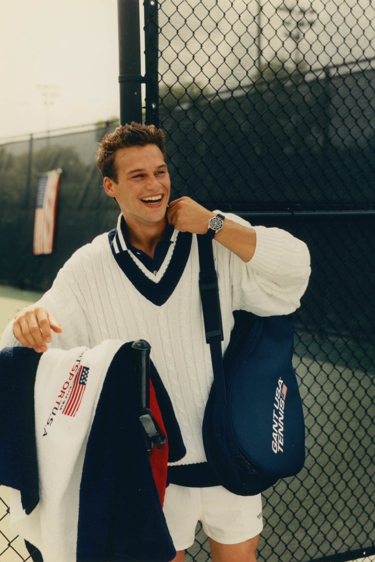 Man wearing a Gant cable‑knit V‑neck sweater with a collared shirt and shorts while holding a tennis bag beside a court fence.