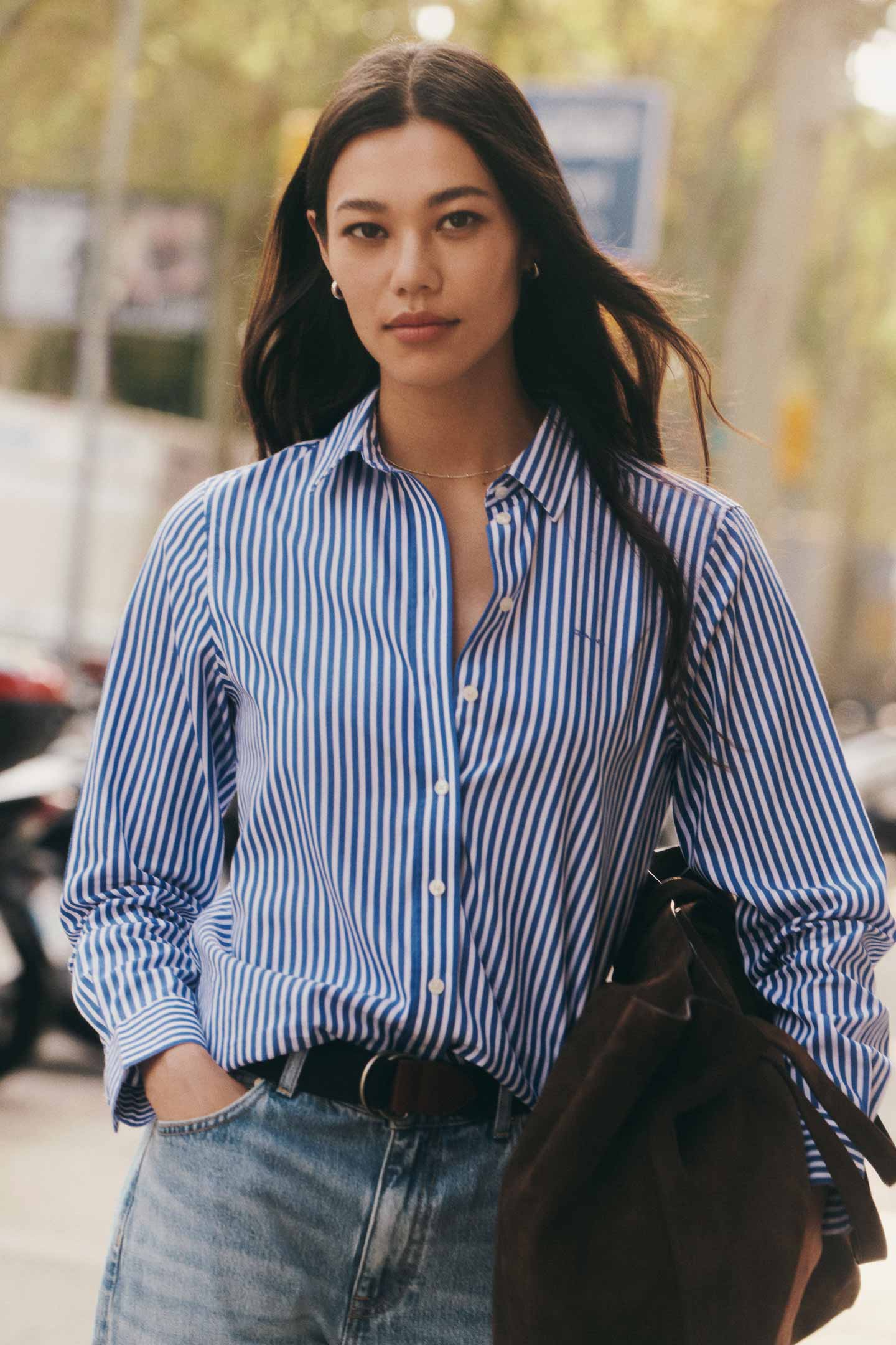 Woman wearing a Gant blue striped shirt with denim jeans while walking outdoors.