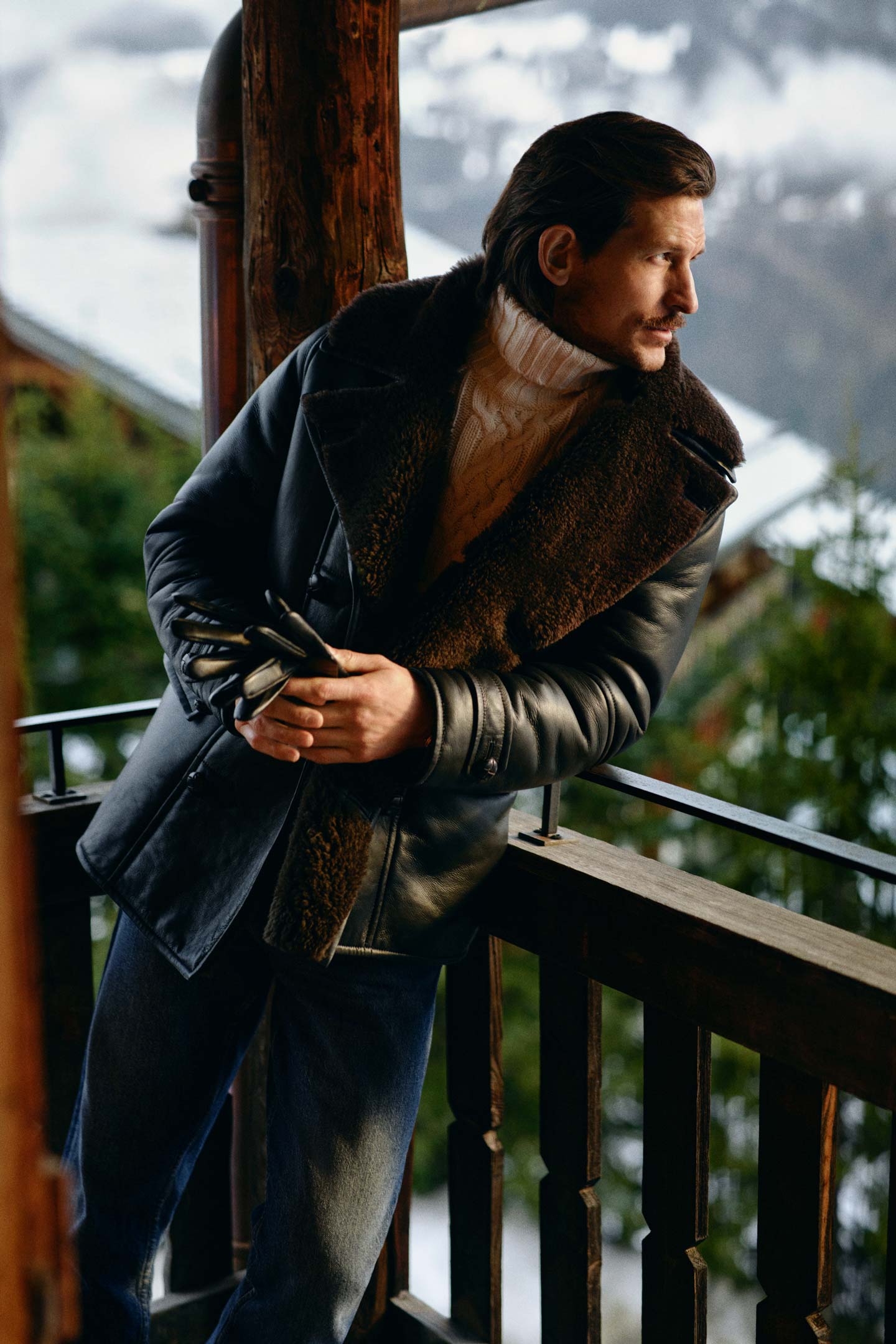 Model styled in a Gant shearling coat layered over a cable-knit roll-neck sweater and jeans on a balcony.