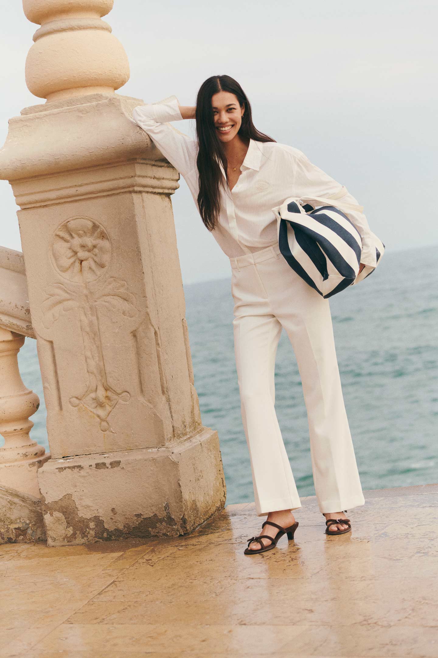 Woman wearing a Gant white shirt and white trousers holding a navy and white stripe canvas beach bag while leaning on a stone railing near the sea.