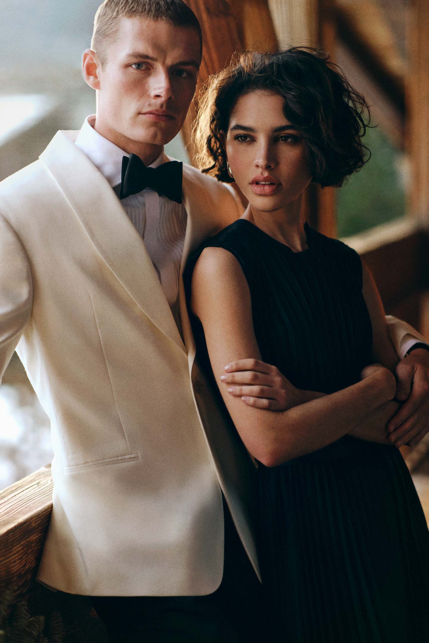 Two models on a balcony, Man styled in a Gant tuxedo jacket with pleated shirt, bow tie and tailored pants; Woman styled in a Gant sleeveless pleated midi dress.