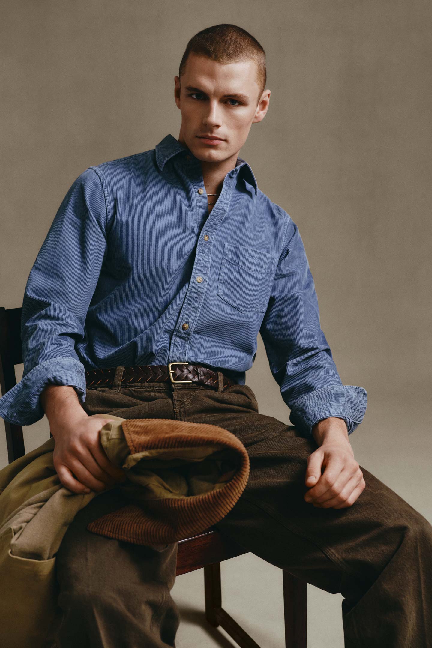 Model styled in a Gant corduroy shirt paired with corduroy pants, seated on a chair holding a folded jacket.