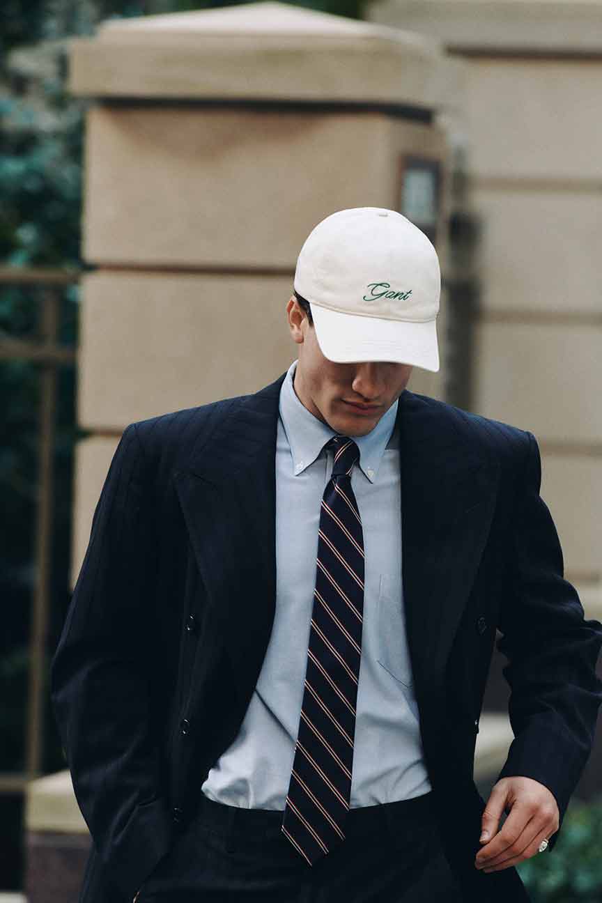 Man wearing a Gant pinstripe suit, shirt, tie and a white Gant cap walking outdoors