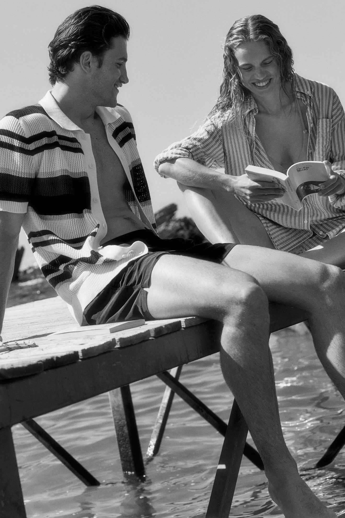 Man and Woman sitting on a pier by the water, Man is wearing Gant knit cardigan and shorts and the Woman is wearing a Gant striped shirt and shorts.
