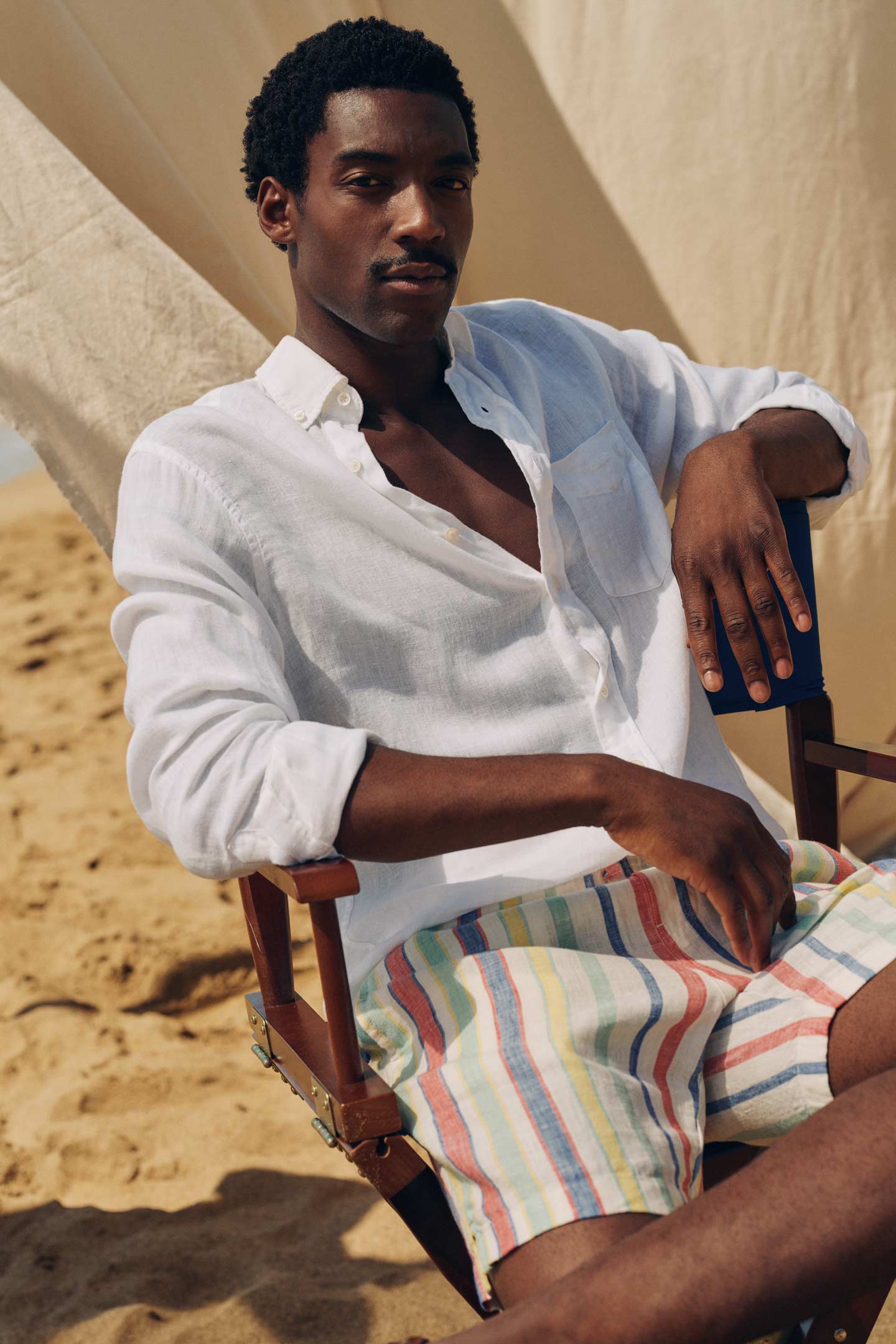 Man wearing a Gant white linen shirt and striped shorts while seated on a beach chair.